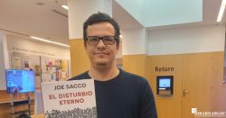 Home dins de la Biblioteca Jaume Fuster sostenint el llibre “El disturbio eterno” de Joe Sacco davant d’un taulell i la màquina de retorn.