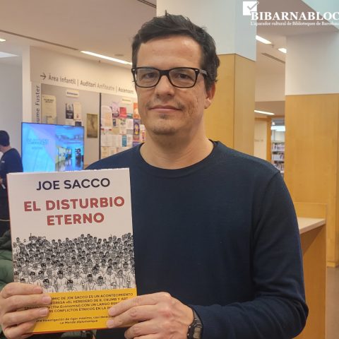 Home dins de la Biblioteca Jaume Fuster sostenint el llibre “El disturbio eterno” de Joe Sacco davant d’un taulell i la màquina de retorn.