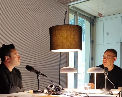 Albert Lladó i Toni Sala conversant davant de micròfons a l'estudi de ràdio de la Biblioteca Gabriel García Márquez amb llums de taula i una làmpada penjant, al costat d’un finestral