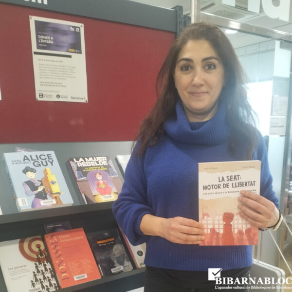 Dona sostenint el llibre “La seat: motor de llibertat” davant d’un prestatge de llibres en una biblioteca.